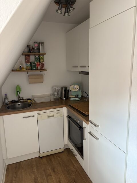 Küche unter der Dachschräge mit Spüle, Holzarbeitsplatte, Backofen, Spülmaschine, Kaffeemaschine, Wasserkocher und einem Regalbrett mit Vorräten