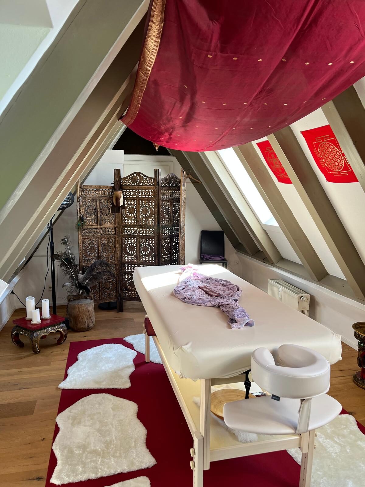 Massageliege unter der Dachschräge, dahinter ein geschnitzter Paravent, Kerzen und ein roter Schirm