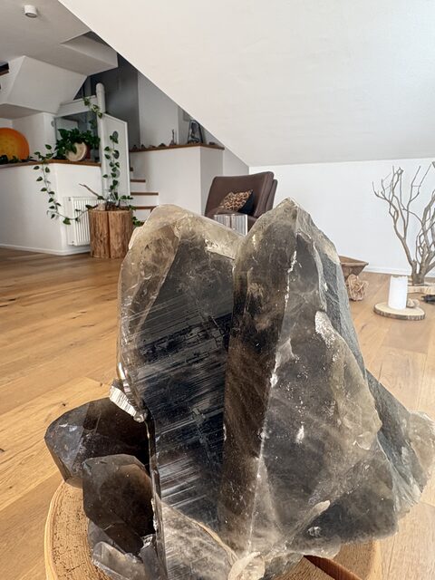 Grosse Rauchquarz-Stufe mit schwarzem Turmalin auf einer Holzscheibe, dahinter der Raum mit Sessel und Treppe
